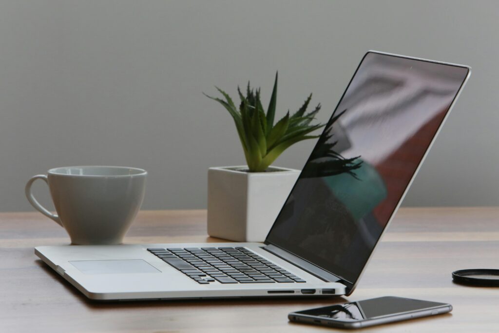 An open laptop, a smartphone, a coffee mug, and a small green plant placed on a wooden table. A white wall is in the background.